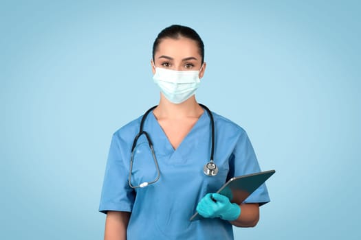 Confident young woman doctor, nurse in blue uniform , protective mask and gloves holding clipboard, blue background
