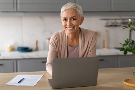 Happy senior woman freelancer working on laptop from home kitchen