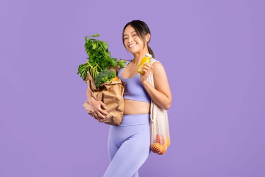 Happy asian woman in sportswear holding bag with veggies and fresh smoothie