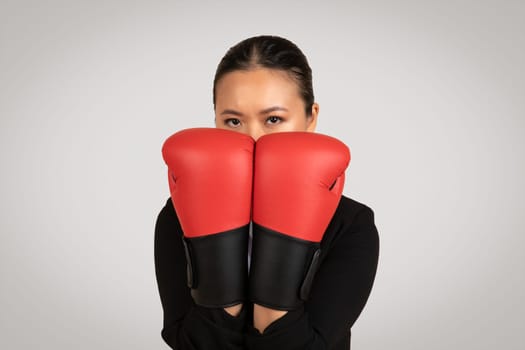 Determined Asian businesswoman with a focused gaze, peeking through red boxing gloves