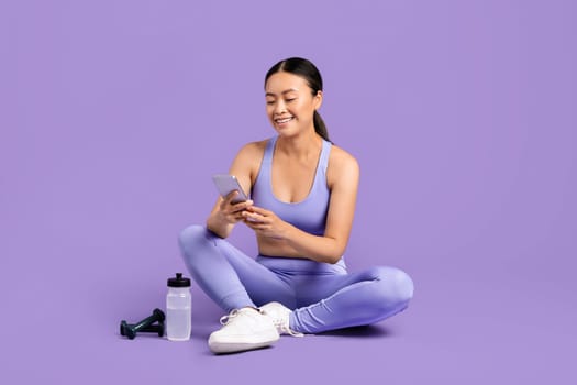 Smiling asian woman using phone during workout break with water bottle