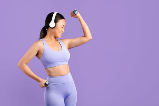 Asian woman in sportswear with headphones flexing muscles with dumbbells