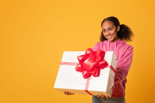 Smiling black youngster girl holds gift box on yellow background