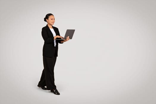 Multitasking Asian businesswoman in a black suit standing and working on a laptop