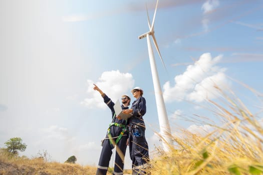 Wide shot professional worker woman hold laptop and stand with her co-worker man point forward and they stand in front of wind turbine or windmill in the field for power plant business.