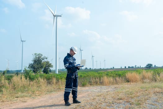 Wide shot of technician worker hold laptop and look to the monitor for working in front of windmill or wind turbine with blue skay in area of power plant factory.