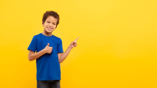 Happy latin school boy giving thumbs up and pointing at copy space on yellow backdrop