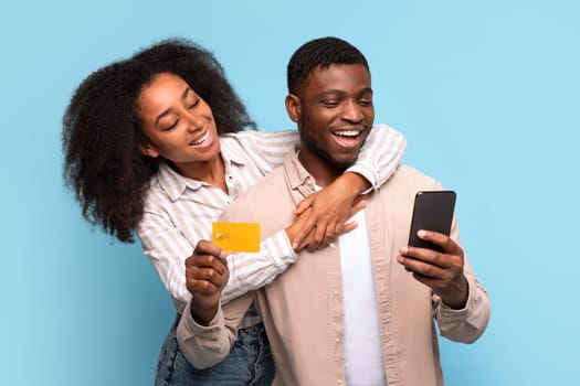 Happy black couple with credit card and smartphone on blue backdrop