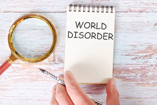 World Disorder written on a blank sheet of a notebook which is touched by a man's hand with a pencil and a magnifying glass is lying next to it