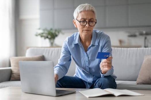 Senior woman shopping on laptop frowning holding credit card indoors