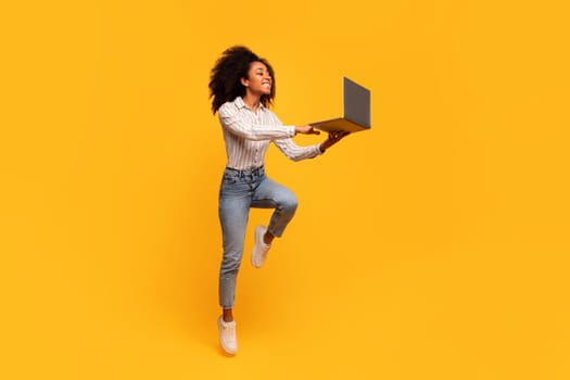 Joyful black woman leaping with laptop on yellow background