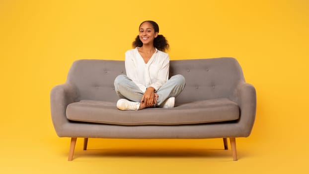 Cheerful african american teen girl sitting on sofa, yellow backdrop