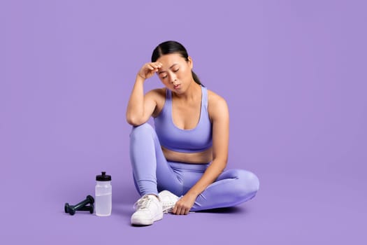 Tired asian woman resting after workout with water bottle and dumbbell