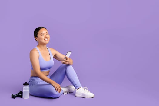 Smiling asian woman enjoying break from fitness, sitting with phone and water bottle, reflecting an active and connected lifestyle on purple backdrop, free space