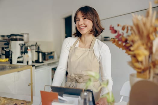 Beautiful female barista is looking away and smiling while standing near bar counter in cafe