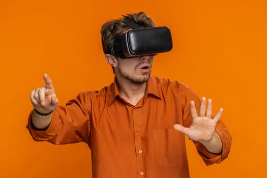Excited bearded man using headset helmet app to play simulation game. Watching virtual reality 3D 360 video imagination. Young guy in VR goggles isolated on orange background indoor. Future technology