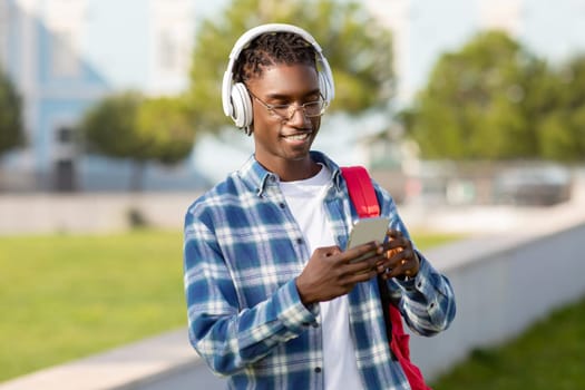 Smiling black student guy with wireless earphones using cellphone outdoors