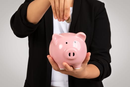 Smartly dressed businesswoman inserting a coin into a pink piggy bank, conceptually illustrating savings