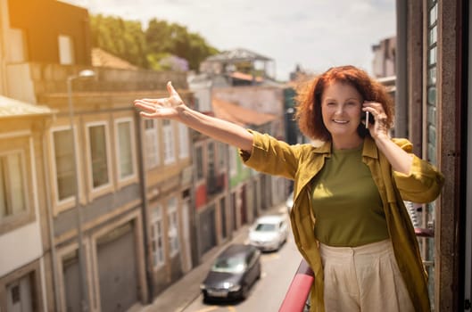 Senior woman standing on sunny balcony, talking on cellphone and waving hand