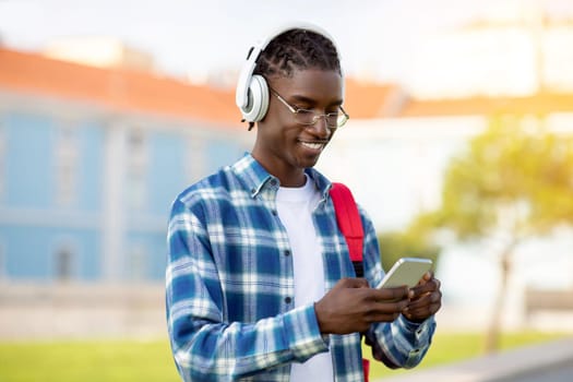 African student guy on cellphone listening audiobooks with earphones outdoors