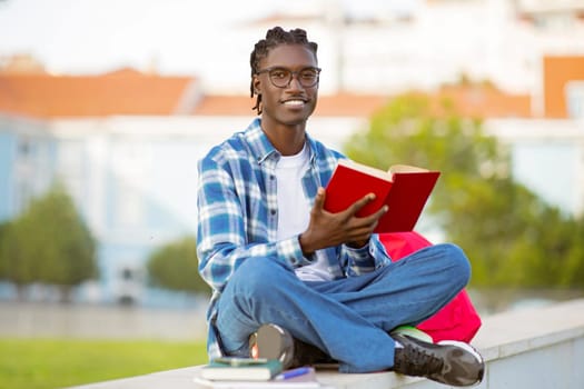Cheerful african american student guy sitting with book learning outside