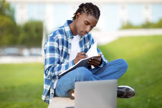Black student guy studies outdoors writing and browsing on laptop
