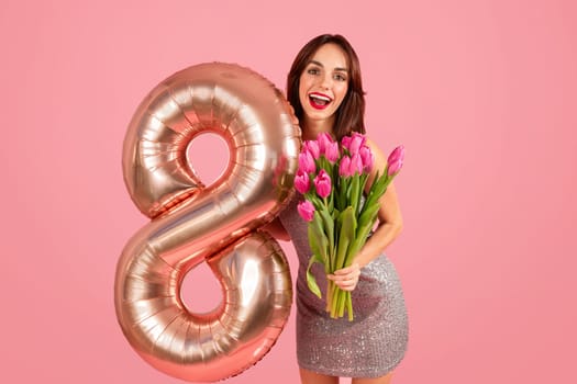 Radiant woman in a sparkling dress joyfully holds a bouquet of pink tulips and a number 8 balloon