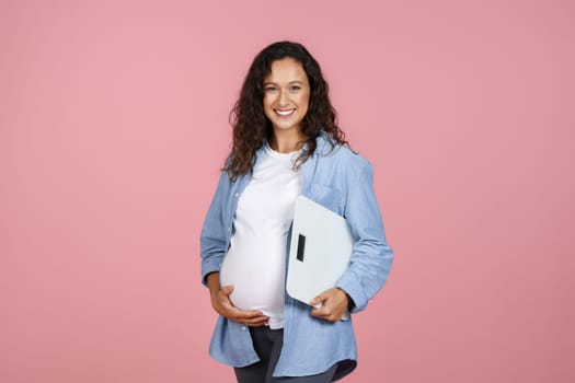 Positive young pregnant woman holding scales, checking weight