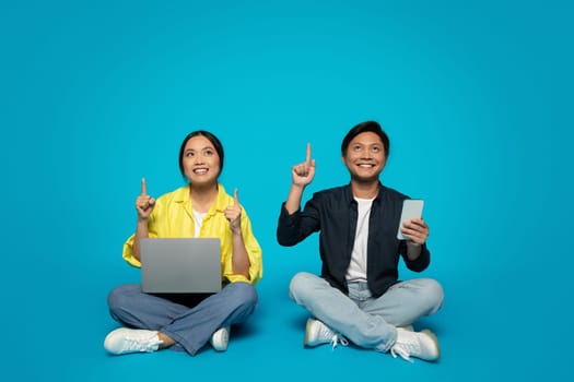 Joyful woman and man sitting with tech devices, pointing upwards, indicating a shared idea