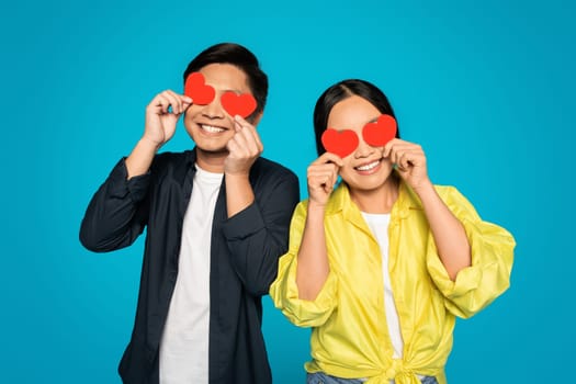 A joyful man and woman playfully cover their eyes with red heart-shaped cutouts