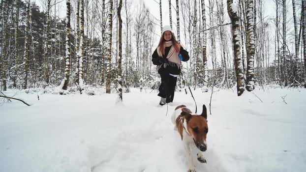 A girl and her Jack Russell Terrier dog are running through the woods.