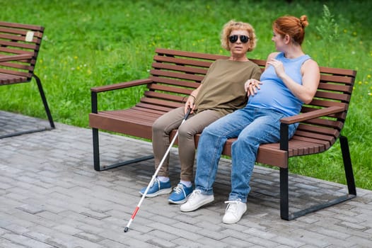 An elderly blind woman and her pregnant daughter are sitting on a bench in the park.