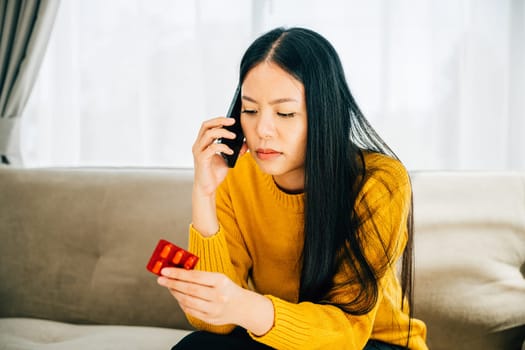 An ill Asian woman sits on a couch taking medicine consulting doctor via phone call at home