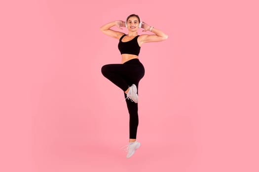 Active young woman wearing headphones jumping against pink studio background