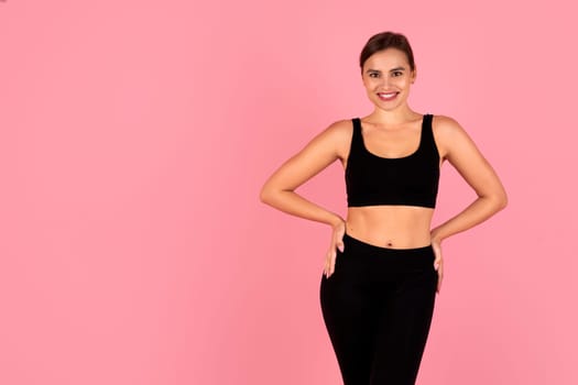 Smiling woman in sportswear with hands on hips posing on pink background