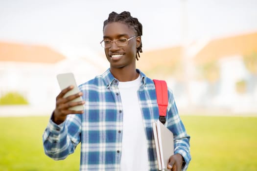 Happy black student holds smartphone, chats and studies online outside