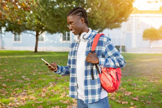 Smiling black student guy browsing on phone with headphones outdoors