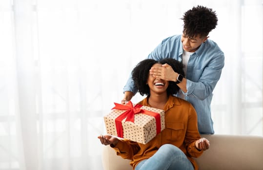 Smiling young african american man closed eyes to lady, gives gift box, in living room interior