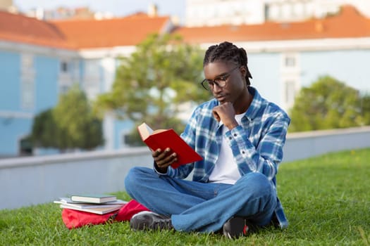 Pensive African American student guy reading book sitting on lawn
