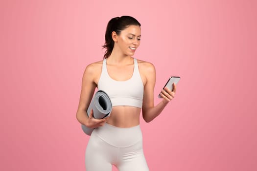Smiling millennial caucasian woman in white sportswear hold mat and chatting on phone