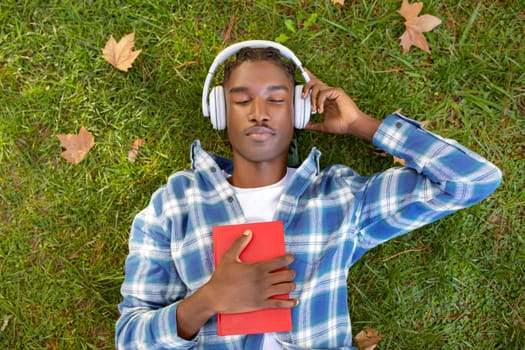 African student guy enjoys audiobook in headphones holding book outdoor