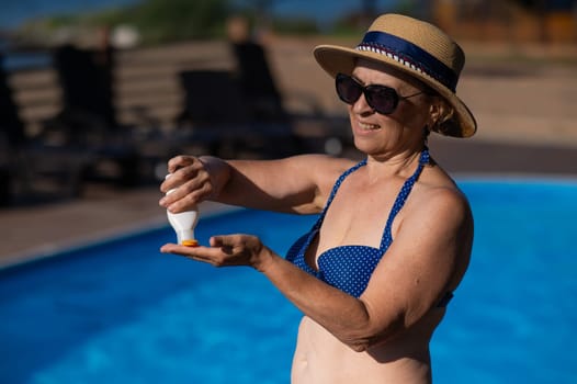 Portrait of an old woman in a straw hat, sunglasses and a swimsuit applying sunscreen to her skin while relaxing by the pool.