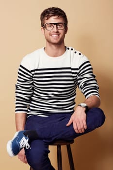 Portrait, fashion and glasses with a man on a chair in studio on tan background for trendy style. Relax, casual and eyewear with a confident young geek or nerd sitting on a stool in a clothes outfit