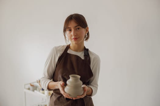 Portrait of young female potter in apron looking at camera while posing in workshop