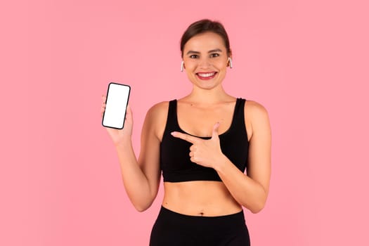 Smiling fitness woman in sportswear joyfully presenting blank smartphone screen