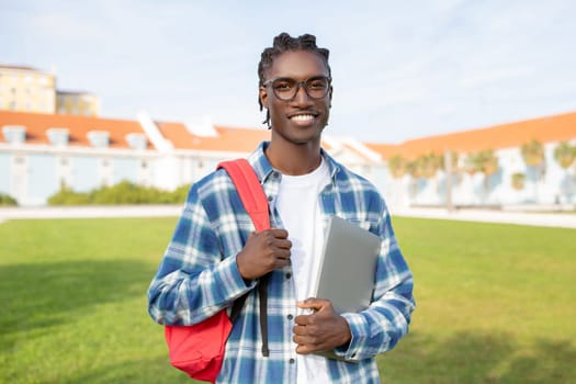 black guy carrying backpack and laptop standing against campus backdrop