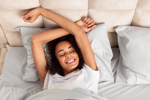 Smiling black millennial woman stretching arms wakes up on bed
