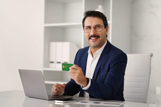 Confident caucasian adult businessman in suit at table with computer, show credit card in modern office interior. Work, business lifestyle, boss recommend finance, investment, saving money