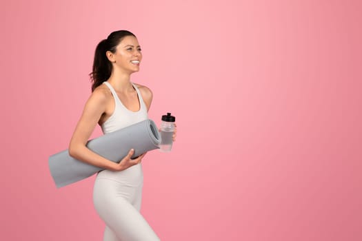Cheerful millennial caucasian lady in white sportswear hold mat and bottle of water