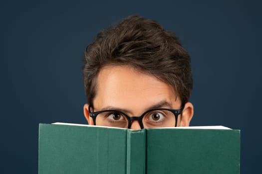 Young man wearing eyeglasses peeking over the top of green book